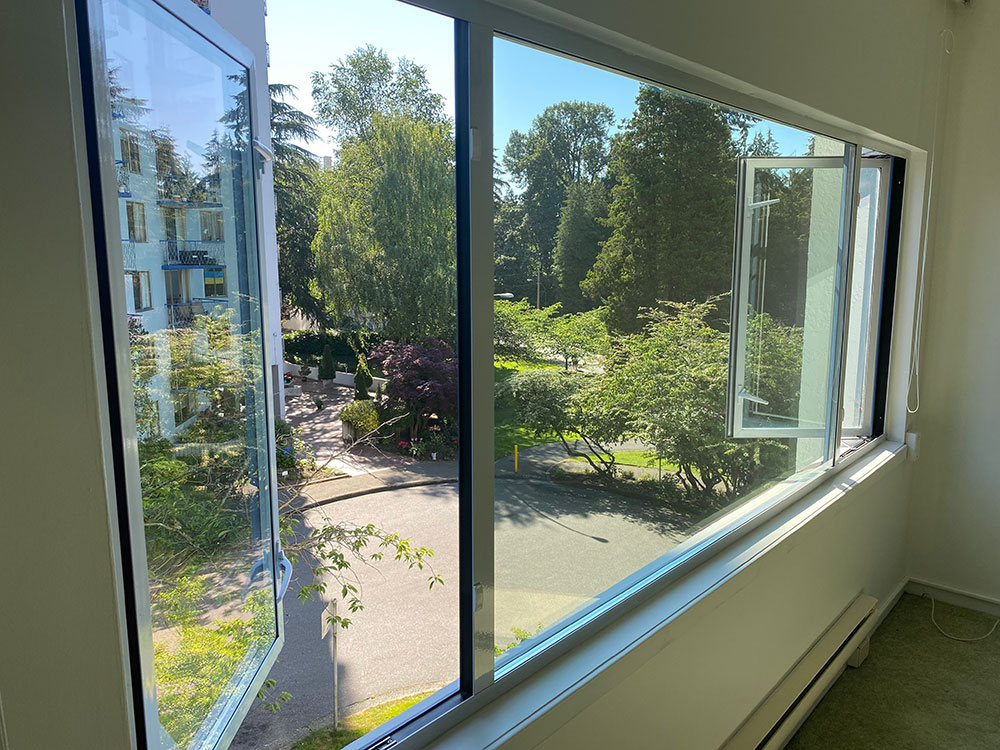 A large second-storey picture window overlooks a sunny street with lush greenery in Vancouver’s West End. Outside, it’s a sunny day and a blue neighbouring apartment building is visible across the street.