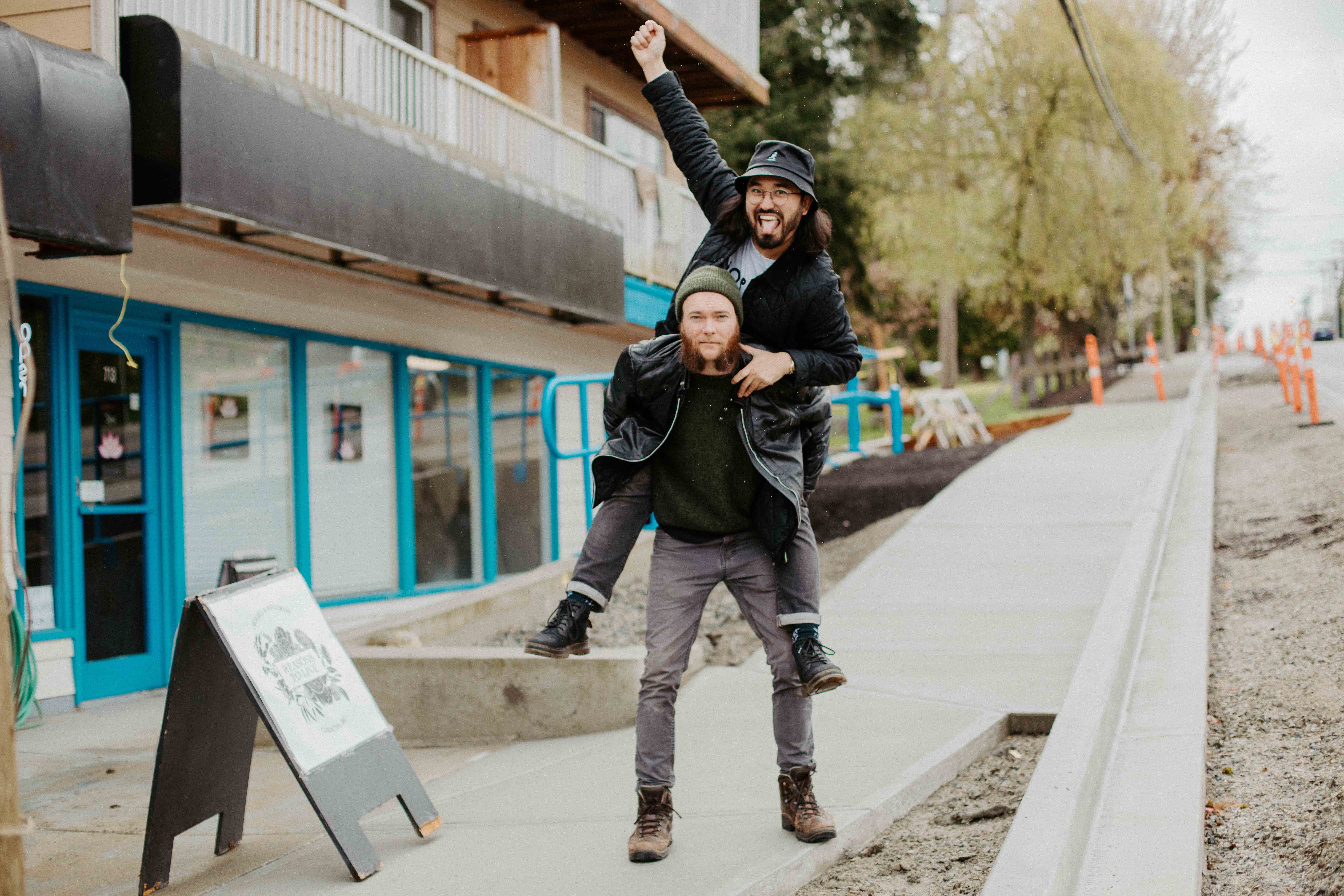 Two young men in black jackets and grey jeans pose on a sidewalk outside a white storefront with turquoise trim. One is carrying the other on his back, who is raising one fist in the air in celebration. He is looking at the camera and smiling with his tongue out.