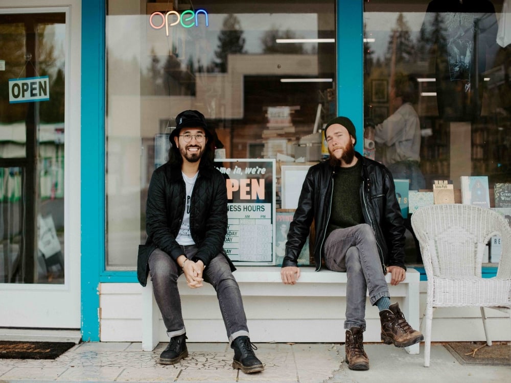 Two men sit on a bench in front of a store. There are various “open” signs pressed against the glass behind them and we can see a person shuffling among the stacks of books in the store.