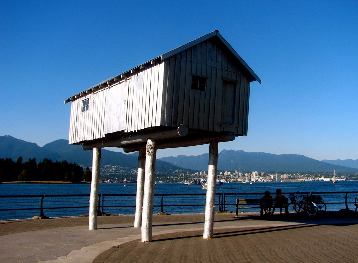 A small white wooden clapboard shed stands on four sideways wooden legs on the Coal Harbour seawall in Vancouver, B.C. The structure perches high above the walkway railing and a nearby bench where people are sitting and facing the water. In the background, the sky and water are bright blue.