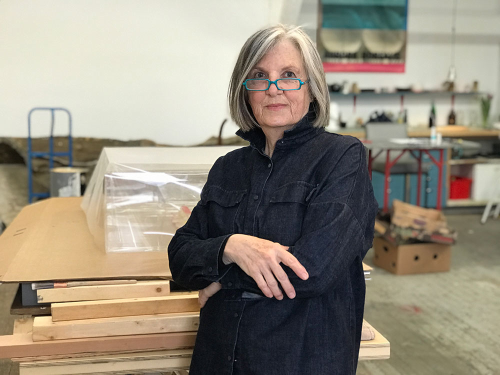 Liz Magor is standing with her arms crossed, looking at the camera with a wise expression. She has short grey hair and a turquoise glasses. She is wearing a dark button-up shirt. Behind her is a stack of unfinished wood in an industrial indoor space, plus boxes of art material.