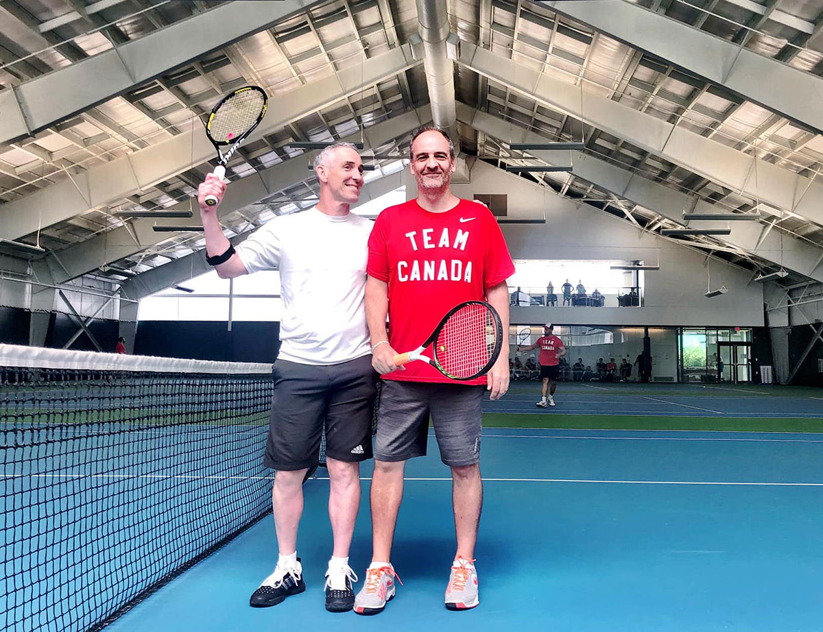 Two men stand together in an indoor tennis court with a blue floor. Jeff Worthington, left, wears a white T-shirt and black athletic shorts; one hand is on the shoulder of his friend while the other holds his tennis racquet aloft. Brian Bella, right, is wearing a red Team Canada t-shirt. Both are smiling.