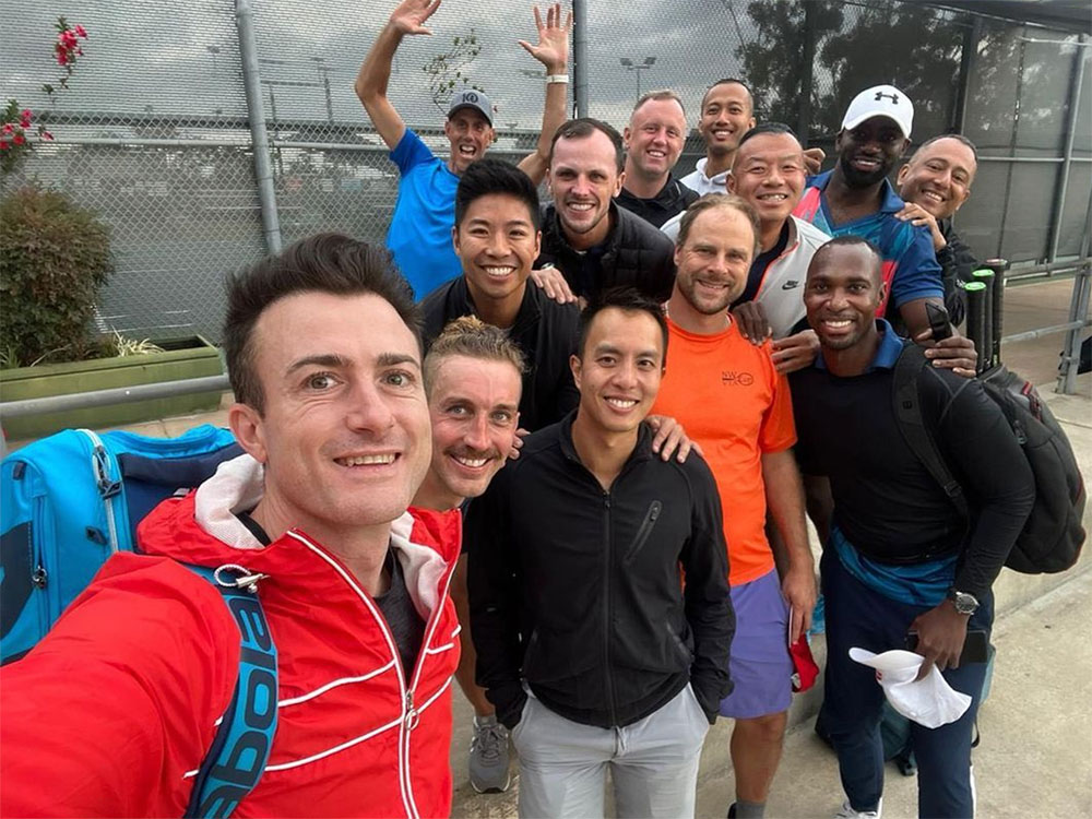A group of 13 men stands closely together, smiling and posing for the camera in front of the gateway to an outdoor tennis court. They are wearing athletic apparel of many colours.