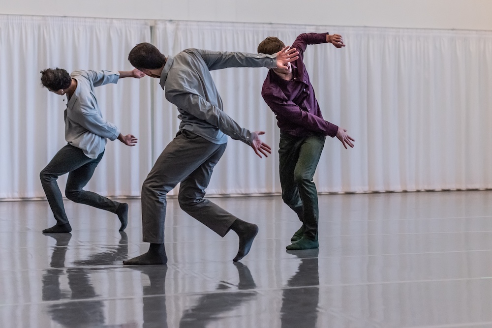 Three dancers in button-down shirts and grey slacks, their faces obscured in the midst of movement.
