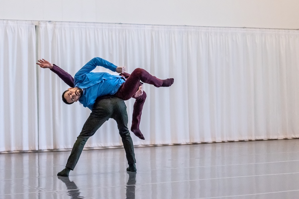 Two dancers, one with their back turned carrying the other, facing the camera in a blue shirt, on their back.