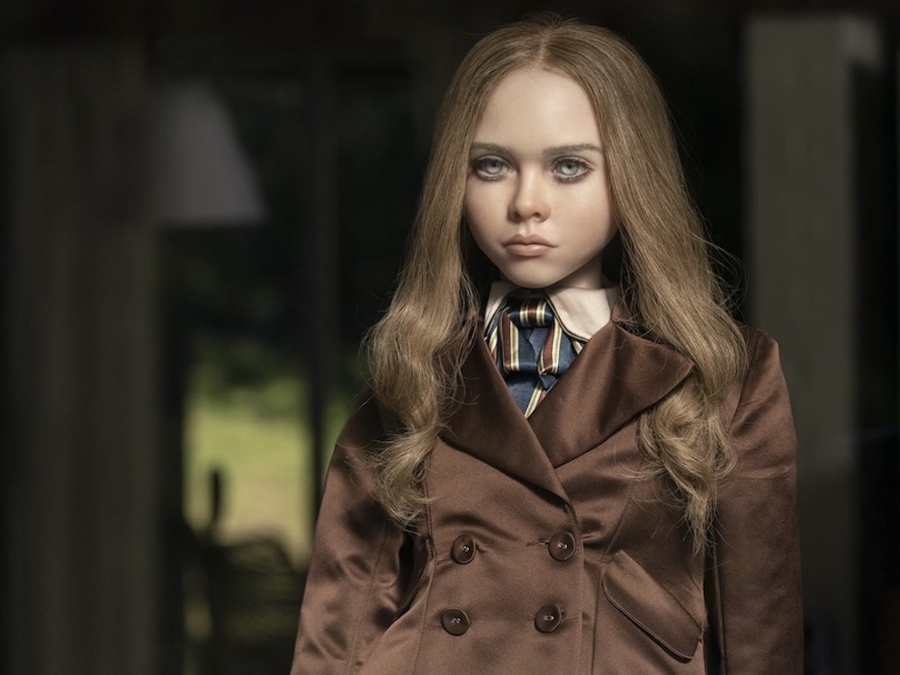 A haunting, hyper-realistic doll wearing a brown silk coat, a cream silk shirt and a striped tie looks blankly into the middle distance.