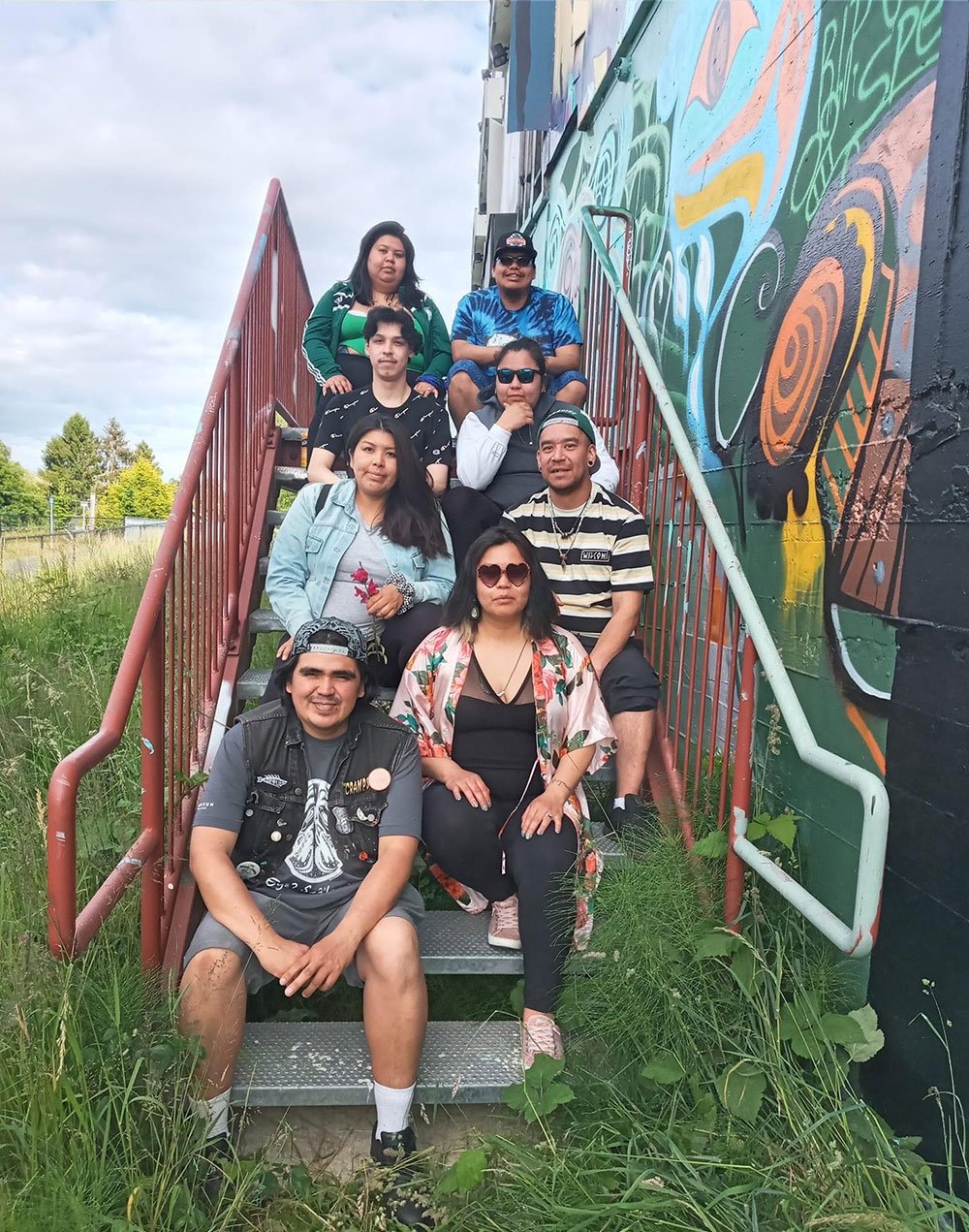 Alex Taylor-McCallum is seated on the bottom stair, to the left of the staircase, with seven people next to one of his pieces of artwork. Long green grass grows around the stairs. It’s a sunny day and people are looking at the camera, smiling.