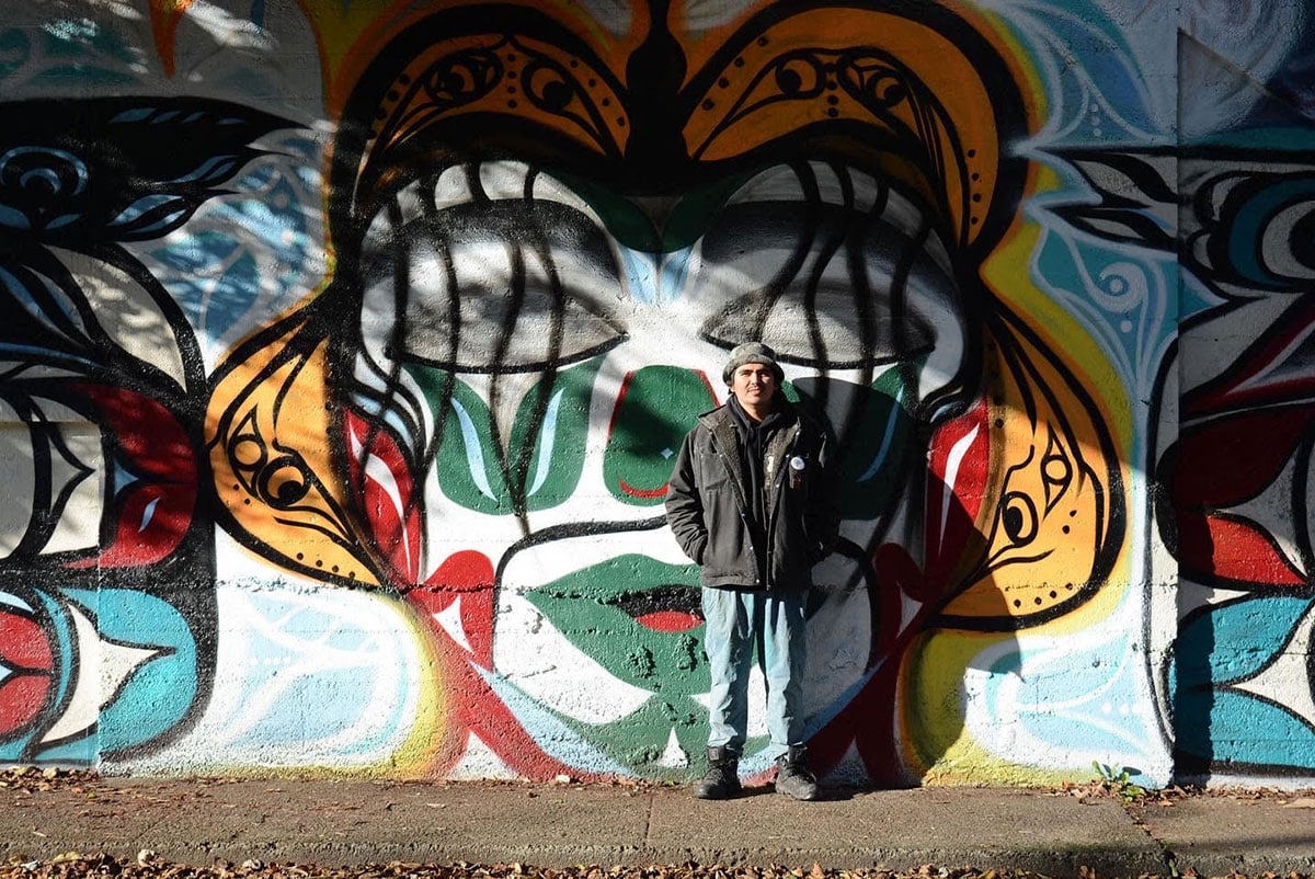 Alex Taylor-McCallum stands in the sun in front of one of his murals in Esquimalt, a stylized illustration of a person’s face. The illustration uses black, orange, white, green and red spray paint. McCallum stands with his hands in the pockets of his black denim jacket. He is wearing a hat and blue jeans.