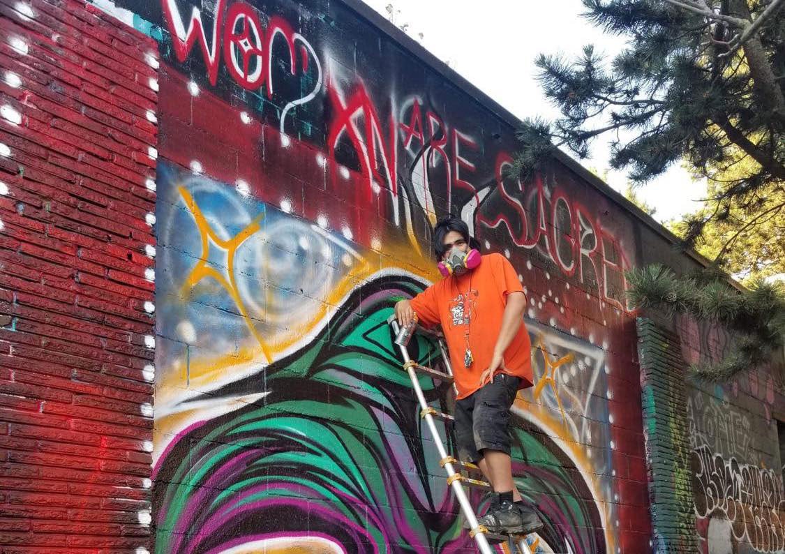 Alex Taylor-McCallum in an orange T-shirt and a respirator at the top of a ladder against a colourful graffiti wall. He is looking down towards the camera and holding a can of spray paint. The wall reads “Womxn are sacred” in handwritten white and red script across the top, below the text is a stylized purple and green illustration of a hummingbird. 