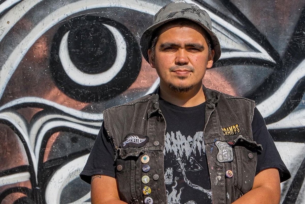 Alex Taylor-McCallum is standing in front of a piece of his public artwork that uses black, grey and bronze paint. Taylor-McCallum is wearing a grey bucket hat and a black denim vest adorned with patches and one-inch buttons over a black band t-shirt with grey punk graphics. He is looking at the camera with a warm, neutral expression.