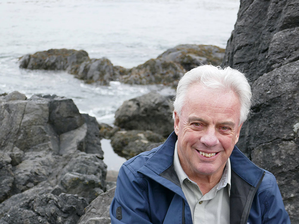A man with grey hair sits or crouches near some rocks on the oceanside, as waves crash behind him.