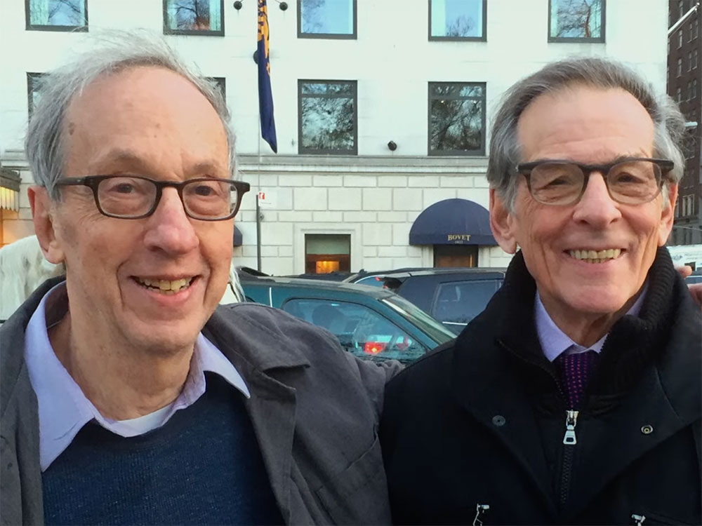 Robert Gottlieb (left) stands with his arm around Robert Caro (right). Both are senior men standing on a busy New York City sidewalk. There is a white horse pulling a handsome cab behind Gottlieb, who is wearing a grey jacket over a navy sweater and light blue collared shirt. Caro is wearing a black pea coat over a light blue shirt and purple tie. Both are wearing glasses and smiling.