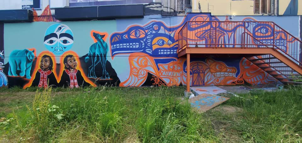 A colourful mural on the side of the Esquimalt Trackside Art Gallery uses blue and orange colourways. The mural is on the side of a one-storey building with an orange staircase.