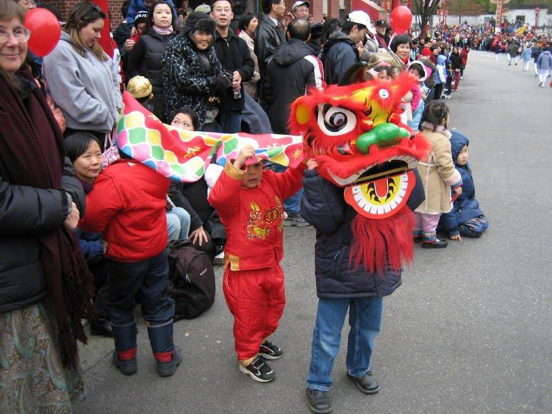 Crowds line the sidewalk leading to Dr. Sun Yat-Sen Garden in Chinatown. To the left of the frame, Fiona Lam is in a black coat, and she is crouching near three small children under a child-sized lion dance costume. Her son is the child in red, holding up the middle of the lion’s body with both hands. 