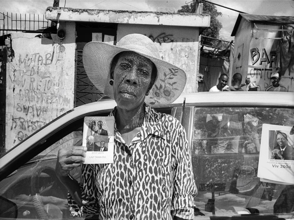 A black-and-white photo depicts a Black woman in a white sun hat wearing a giraffe-patterned top. She is frowning at the camera and holding up a photograph to signal her support of Jean-Bertrand Aristide, Haiti’s first democratically-elected president.