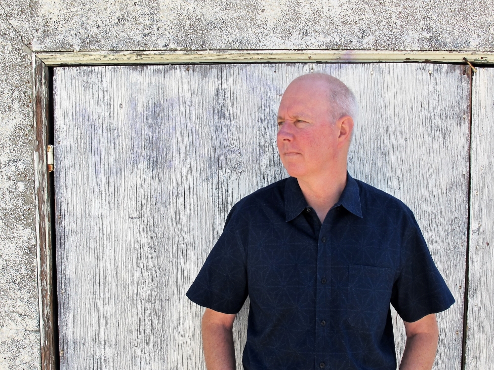 Dave stands in front of a white stucco shed that has weathered wooden doors, also painted white. He looks off to his right.