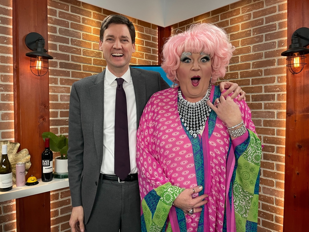 Premier David Eby stands to the left of the frame with his arm around Connie Smudge to the right. Eby is wearing a grey suit, white shirt and burgundy tie. He has short brown hair, is looking at the camera and smiling widely. Smudge is wearing a pink wig and a dress with pink, blue and green patterned layers. She is looking at the camera with an expression of mock surprise. 