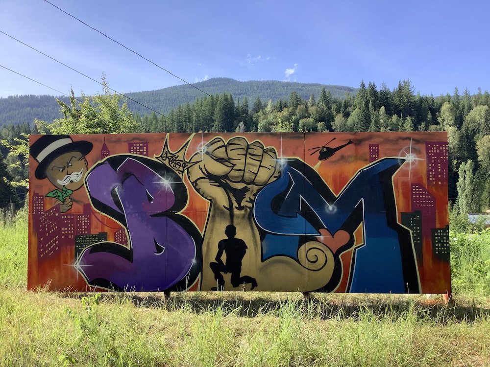 A billboard has been spray-painted. The monocle man from Monopoly holds cash in the top left corner; a police helicopter buzzes above the silhouette of a city. A man kneels in the centre of the mural. The letters “BLM” take up a large amount of space.
