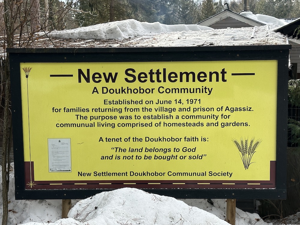 A yellow and black sign reads, “New Settlement: A Doukhobor Community. Established on June 14, 1971, for families returning from the village and prison of Agassiz. The purpose was to establish a communal living comprised of homesteads and gardens. A tenet of the Doukhobor faith is: ‘The land belongs to God, and is not to be bought or sold.’ New Settlement Doukhobor Communal Society.”