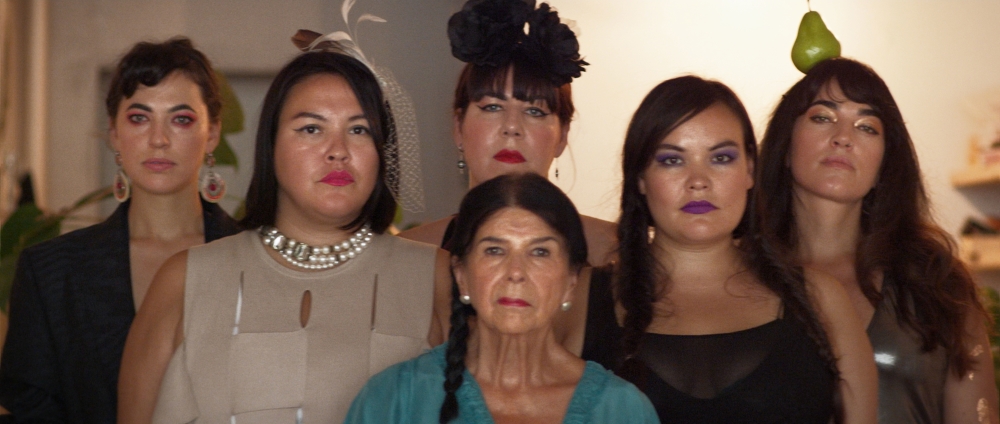 Six women stand together in a dimly lit indoor space. They are wearing makeup and eveningwear. They are looking towards the camera with neutral expressions on their faces.