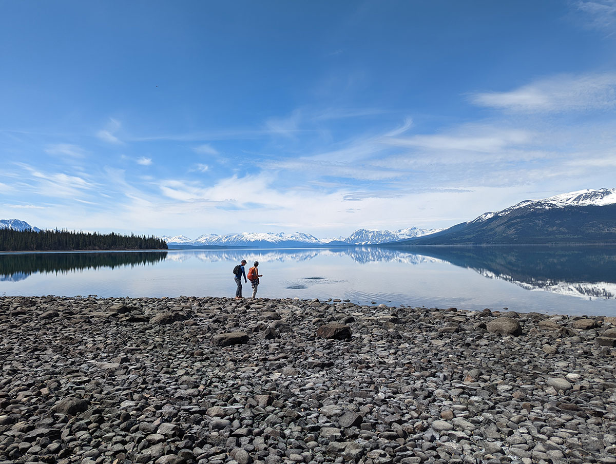 Two figures stand at the edge of a clear lake, skipping stones. Snow-capped mountains are visible in the distance.