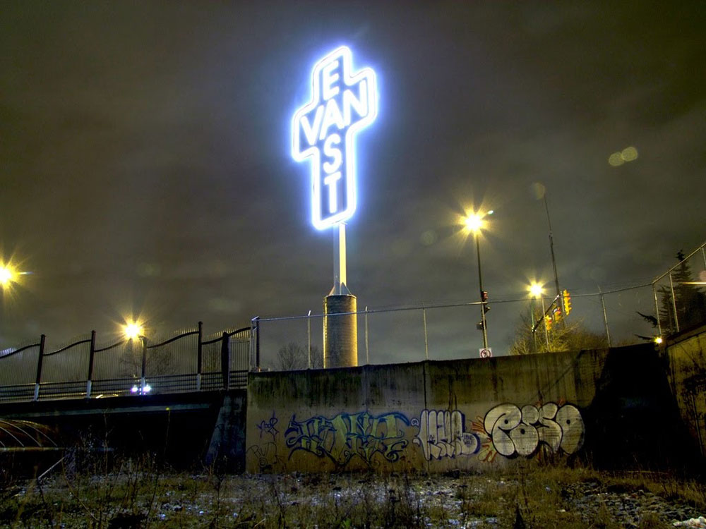 At night, a giant light-up cross rises from the top of an industrial landscape with graffiti. Inside the gross are the letters “East” and “Van,” sharing the letter A.