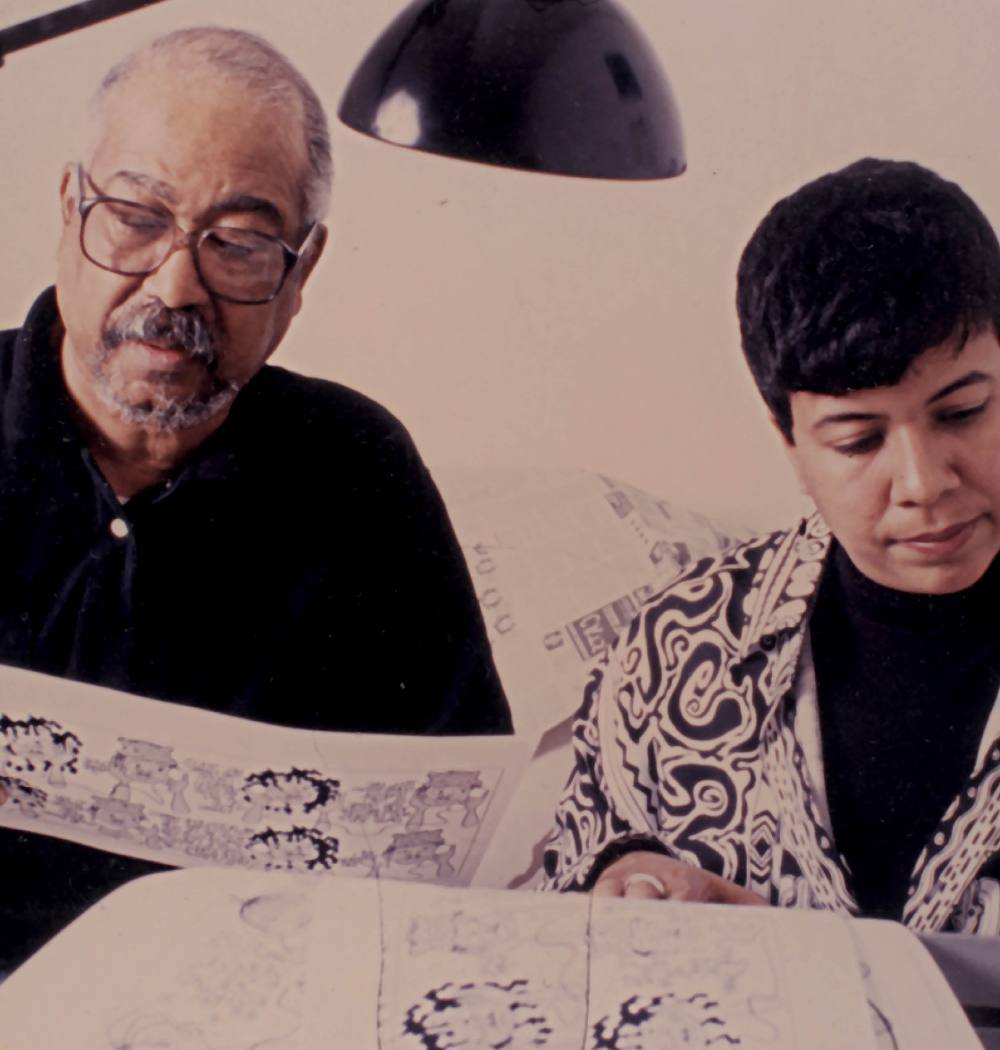 An undated colour photo depicts Brumsic Brandon Jr., left, and Barbara Brandon-Croft. Brandon is wearing glasses and a black golf shirt. Brandon-Croft has short dark hair and is wearing a black patterned shirt over a black turtleneck. They are looking at black and white comic strips together. 