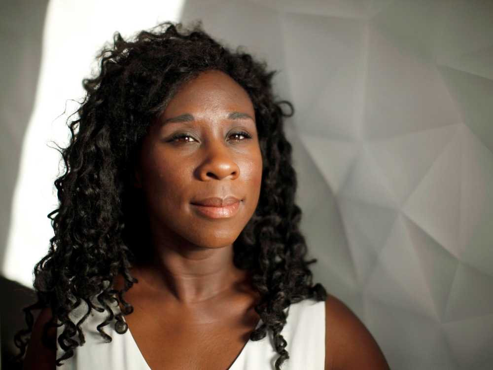 Esi Edugyan is a Black woman looking towards the camera, smiling softly. She has dark curly hair and is wearing white. She is standing against a white background and a piece of white furnishing with geometric faceted lines.  