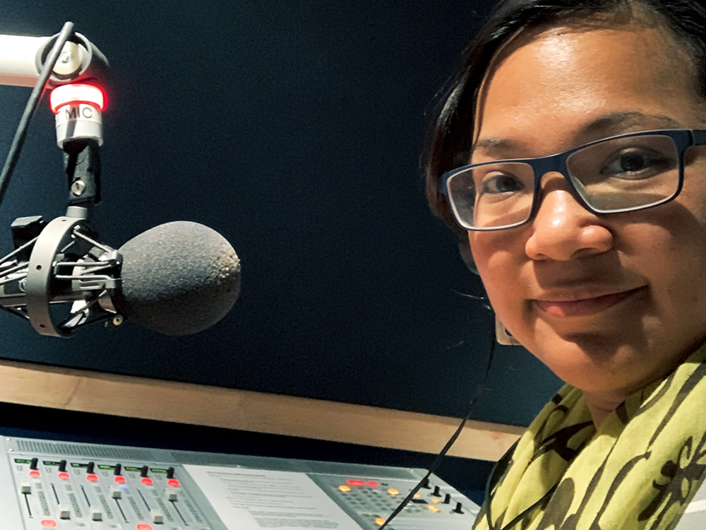 Ethel Tungohan smiles at the camera. She is wearing headphones and sitting in front of a microphone and mixing panel.