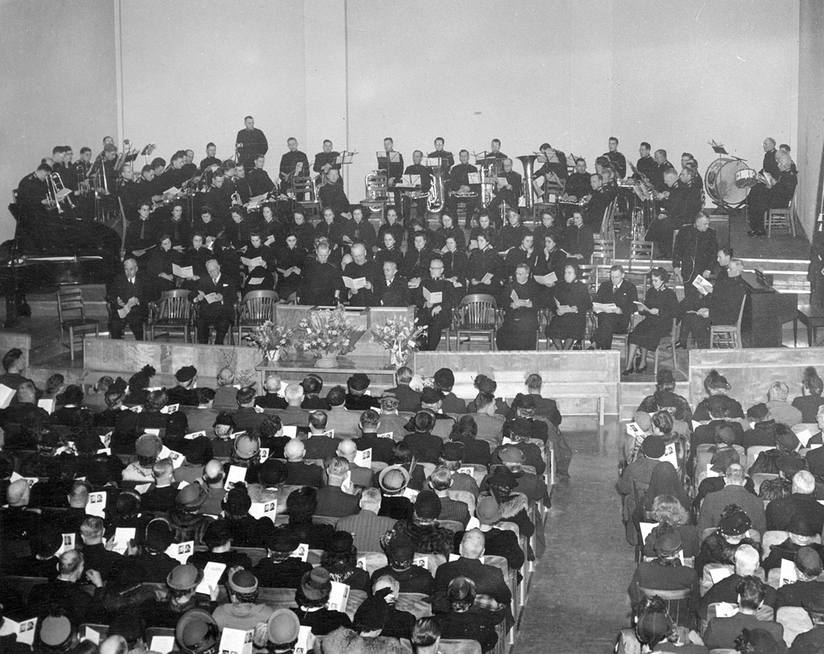 A black and white photo shows a big band playing on stage, from the audience’s point of view. It’s a packed house.