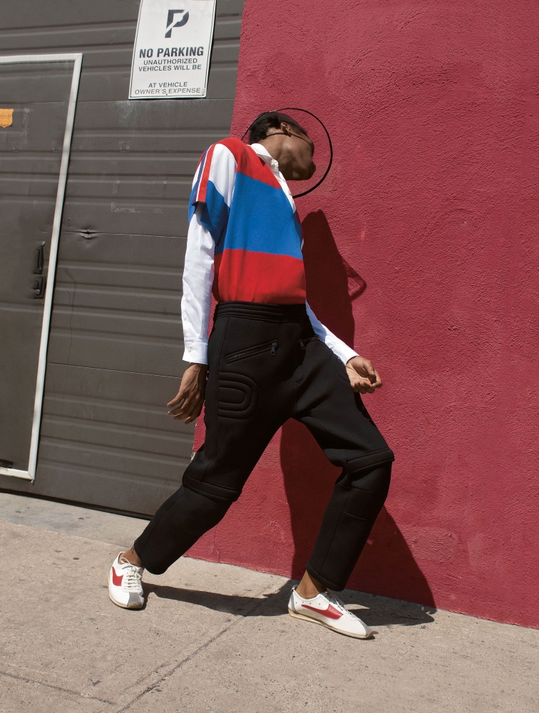 A young black man leans towards the red wall behind him as he walks down a sidewalk. He is wearing a white, blue and red polo shirt, black pants and white sneakers with red stripes.