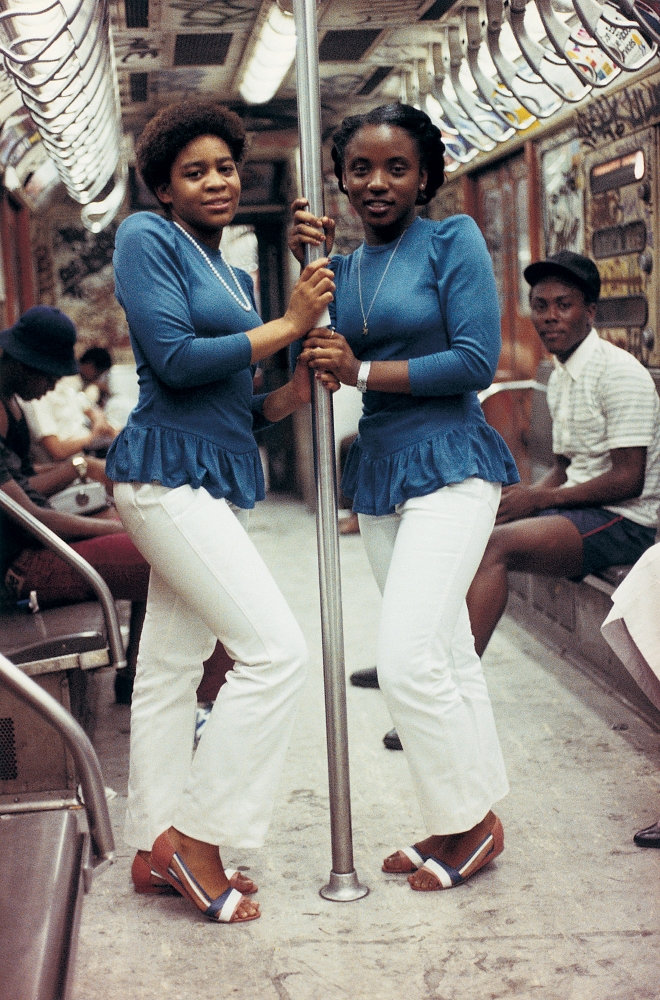 Two Black women in matching outfits stand holding a pole inside a New York City subway car. They are wearing white pants and blue tops with a fringe around the waist.