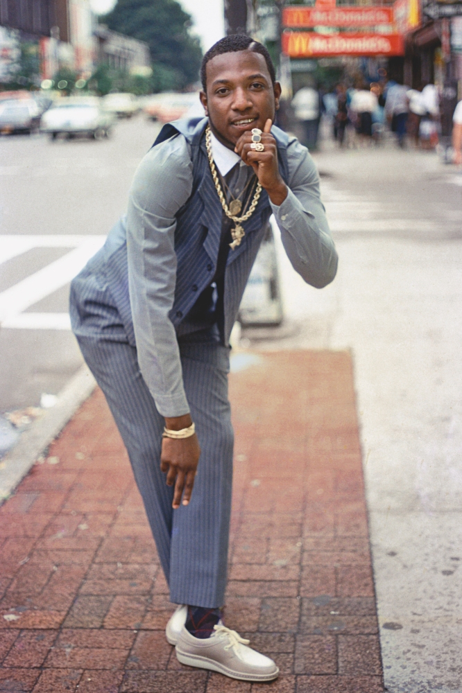 A 1980s colour photograph of a young man in grey pinstripe pants, vest and grey and white lace-up shoes leans towards the camera. He has a gold chain around his neck and several rings on his finger. In the background, a Brooklyn streetscape is out of focus.