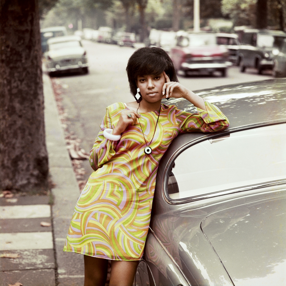 A young Black woman in a long-sleeved dress in a swirling orange, yellow and green ‘60s motif leans against a grey car on a quiet residential street. 