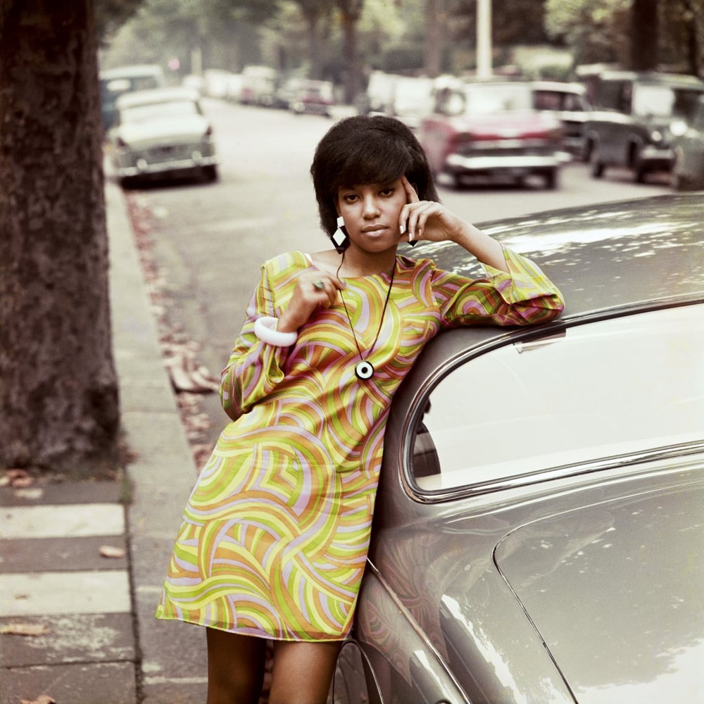 A young Black woman in a long-sleeved dress in a swirling orange, yellow and green ‘60s motif leans against a grey car on a quiet residential street. 