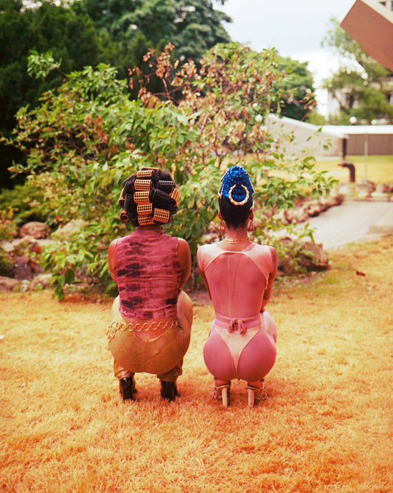 Two Black women are crouching next to each other with their backs turned to the camera. The person on the left has large gold curlers in her hair; the person on the right has an ornamental blue headpiece on with a gold ring affixed to the back. They are wearing shades of pink and gold.