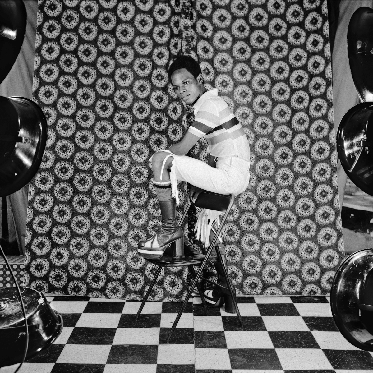 A black and white portrait of a young Black person with short hair, a polo T-shirt, disco boots and fringe shorts posing on a plastic chair. They are standing against a ‘70s patterned wallpaper with circular designs.