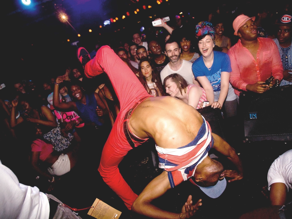 A person in bright red pants and sneakers is bending over backwards into a handstand at a crowded nightclub venue. Their midriff is visible in the foreground. Behind them are crowds of partygoers in colourful streetwear, cheering them on.