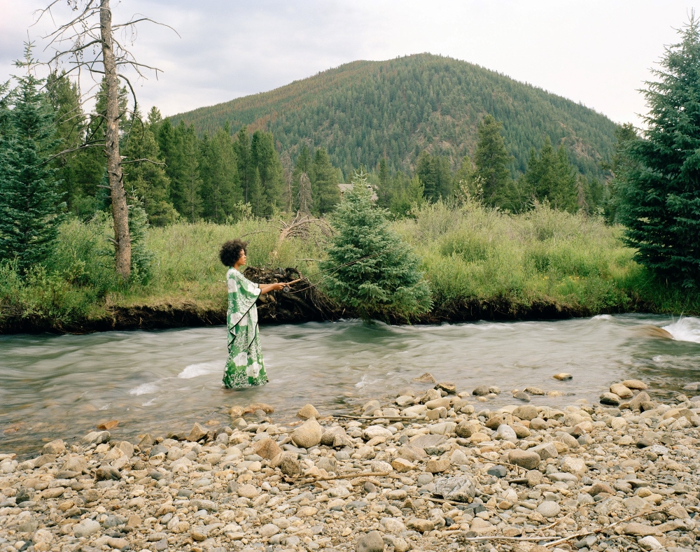  A Black woman stands in the middle of a river against the backdrop of a mountain and forest. She is wearing a flowing bright green and white dress. She is flyfishing.