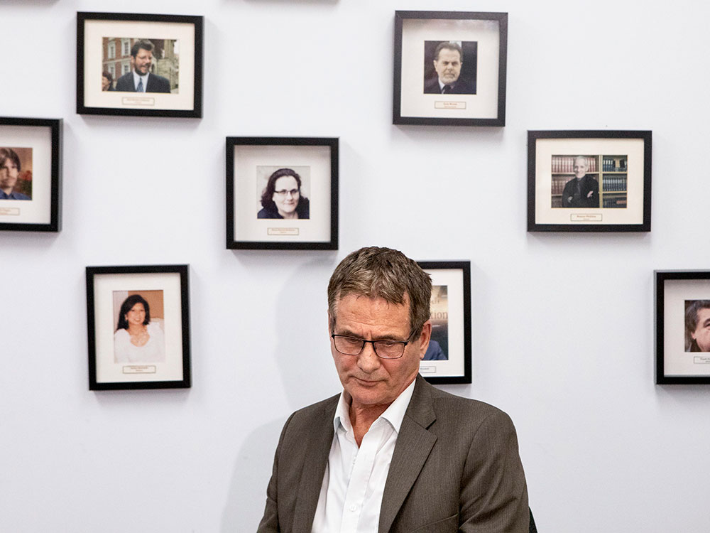 A white man in his late middle age (David Milgaard) stands in front of a white wall with framed photo portraits of other people. 