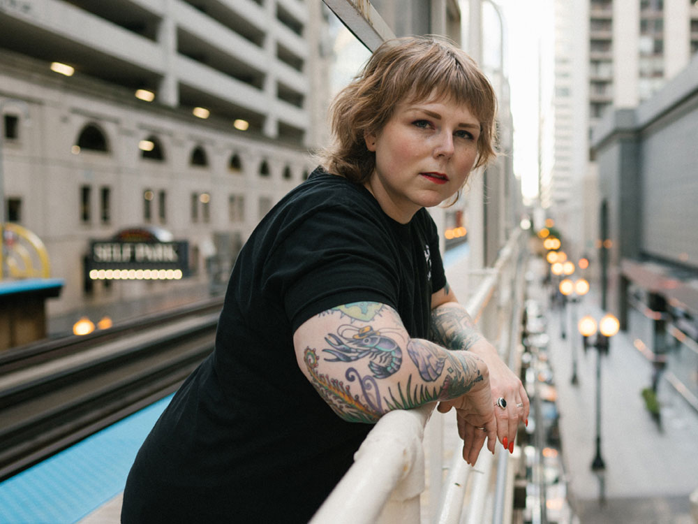Tracey Lindeman leans over the railing of a platform in a busy city. Her forearms are tattooed. She looks at the camera with a level gaze.