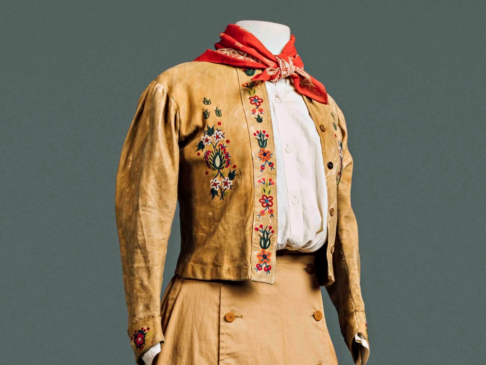 A white mannequin wears a light brown buckskin jacket featuring bright floral embroidery. The mannequin is also wearing a red kerchief around its neck and a long light brown skirt. The mannequin is photographed against a grey background.