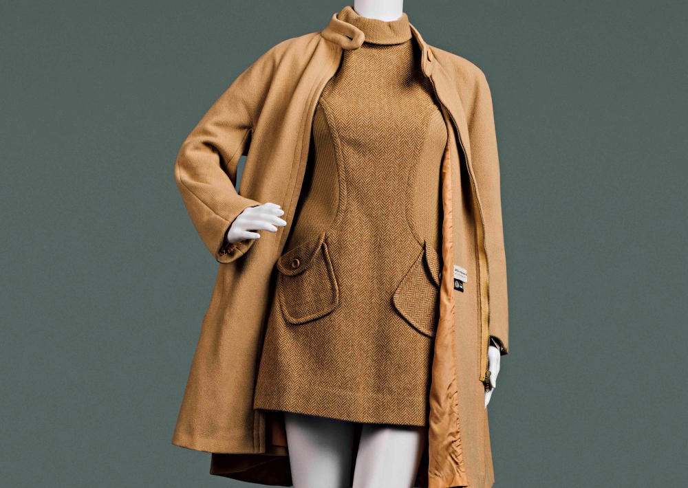 A camel-coloured short dress is worn under a long jacket in a matching colour in a style popularized in the mid-1960s. This detail of the clothing on a white mannequin against a grey background shows the body of the dress and jacket from shoulders to mid-thigh.