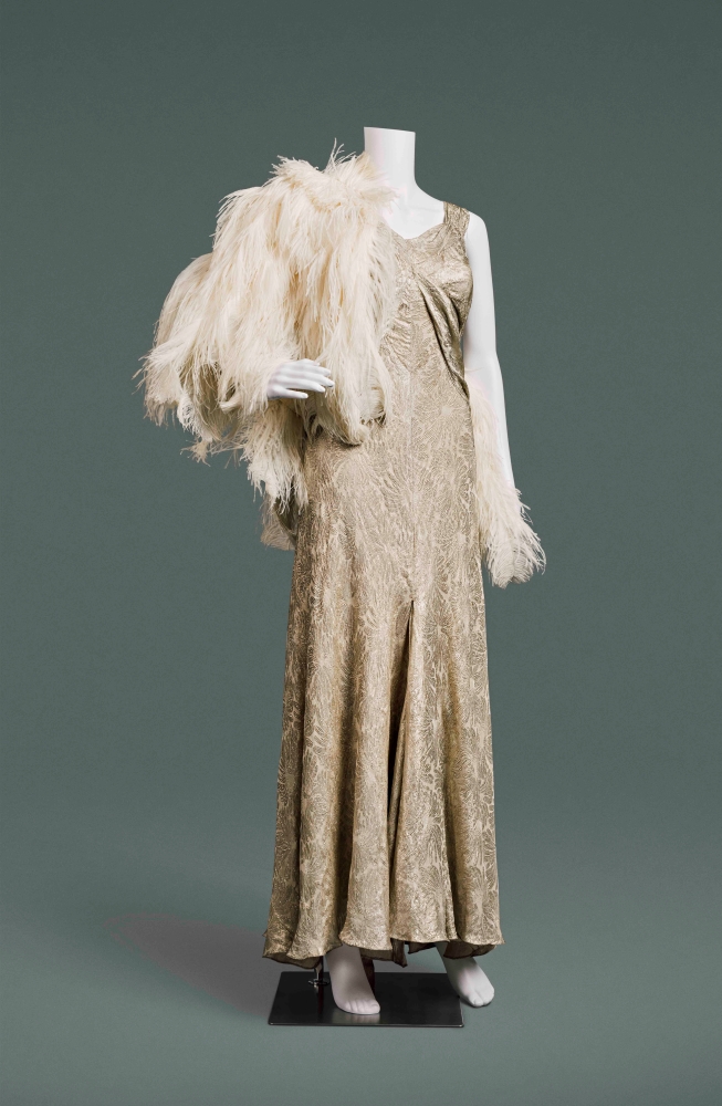 A silver and cream brocade evening dress is on a white mannequin holding a cream feather boa against a grey background.