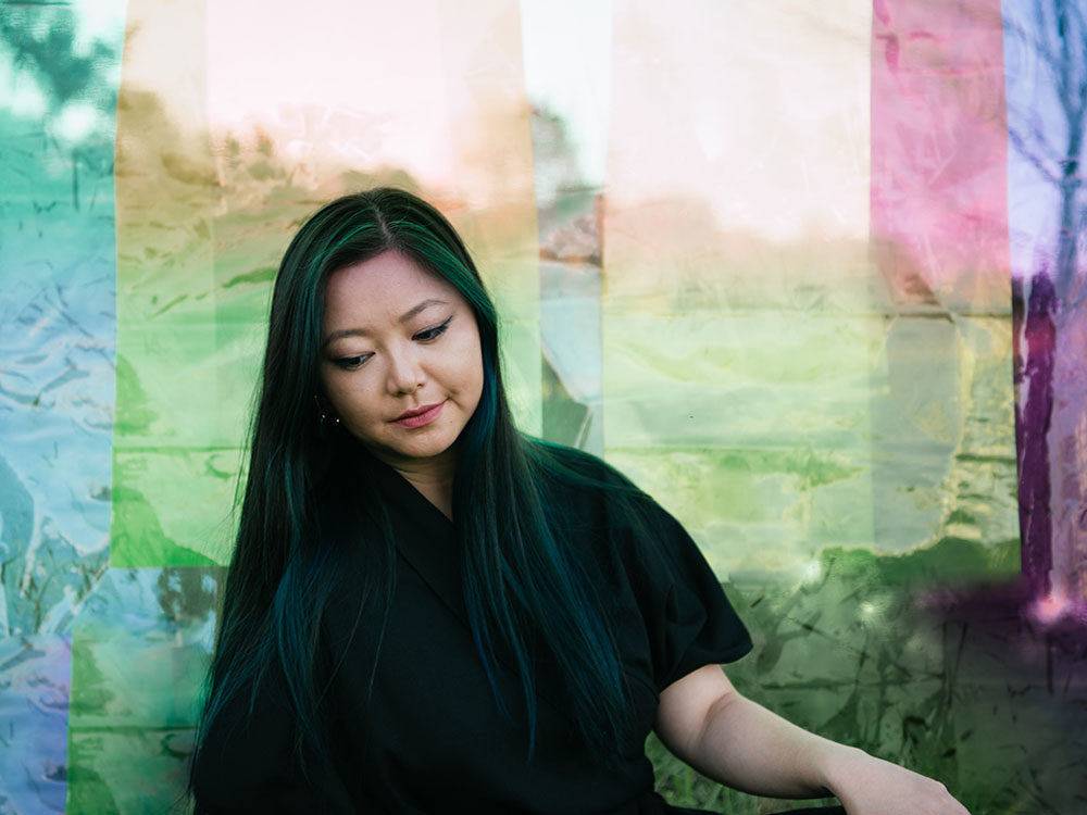 Vicky Chow is a Chinese woman with long black hair and turquoise highlights. She is looking down towards the left of the frame, wearing a black top. She is standing against a transluscent background with layered rainbow colours. 