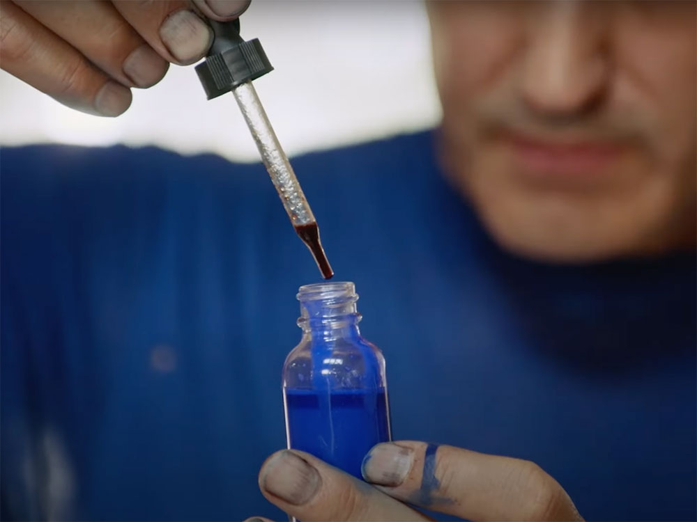 A closeup photo of a man using a dropper to add a drop of dark red ink to a bottle of blue ink. He is wearing a blue shirt the lower half of his face and shoulders are in soft focus in the background. 