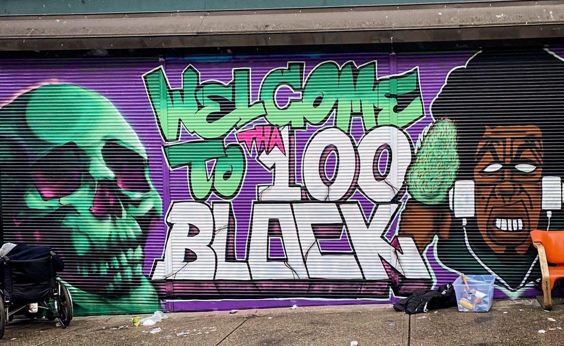 Purple and green spray paint says “Welcome to the 100 block” and depicts a green skull on the left and a Black woman’s face on the right.