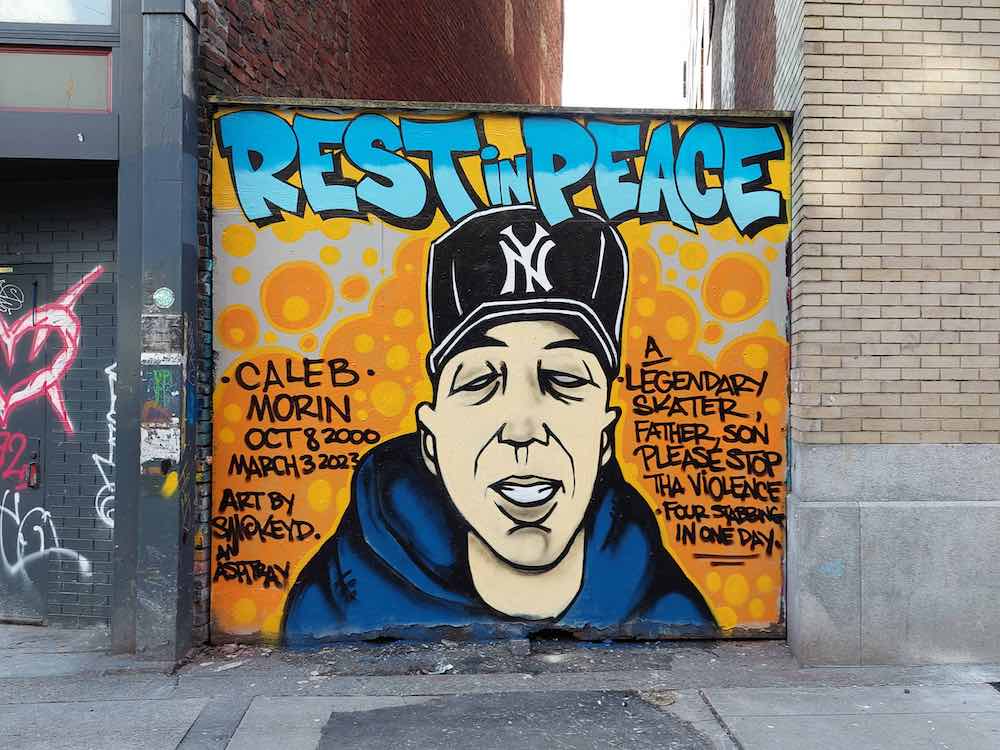 A large-format mural in memory of Caleb Morin features a portrait of Morin from the shoulders up in a blue hoodie and black New York Yankees ball cap. Morin is drawn against a yellow background and a handwritten message dedicated to his memory.