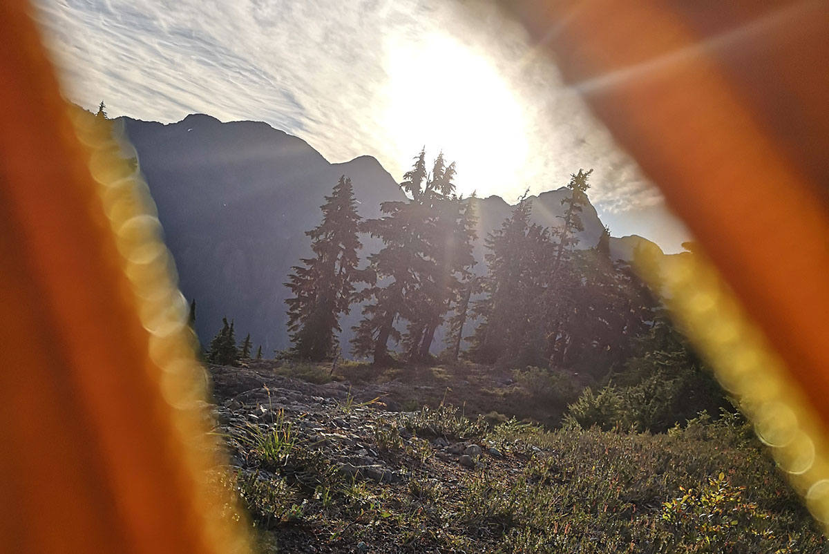 The foreground is rocky, with small trees and some water collecting. The background features the sun rising over the mountains.