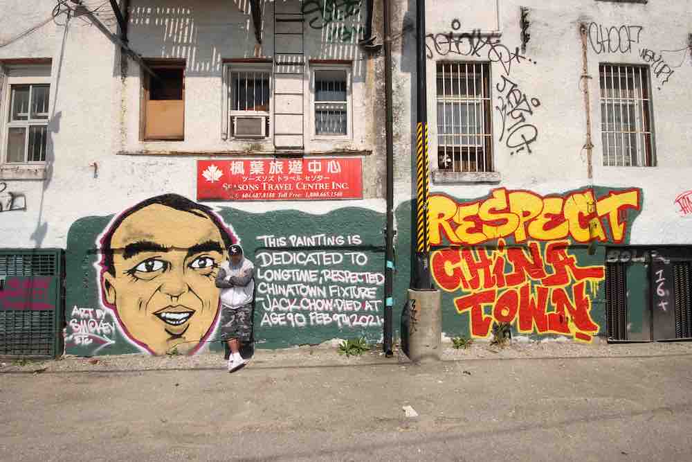 Smokey Devil leans against a muralled wall in a white hoodie and green camo shorts. The mural is wide across a wall, using a forest green background, an illustration of the late Jack Chow’s face, and a message in yellow and read that says “Respect Chinatown.”